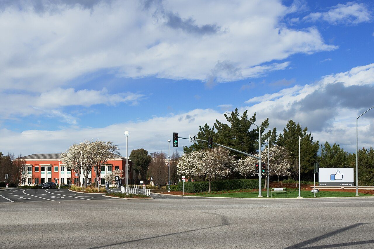 A road junction with traffic lights outside a red-brick office building, with a white sign showing a blue thumbs-up and the address 1 Hacker Way