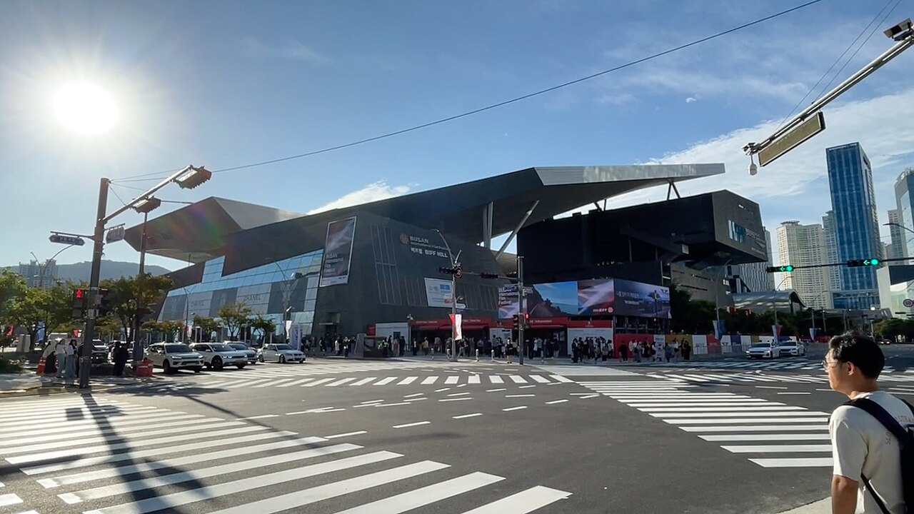 The Busan Cinema Center, a dark building with a huge sloping roof, behind a wide crosswalk with traffic and pedestrians on a sunny day