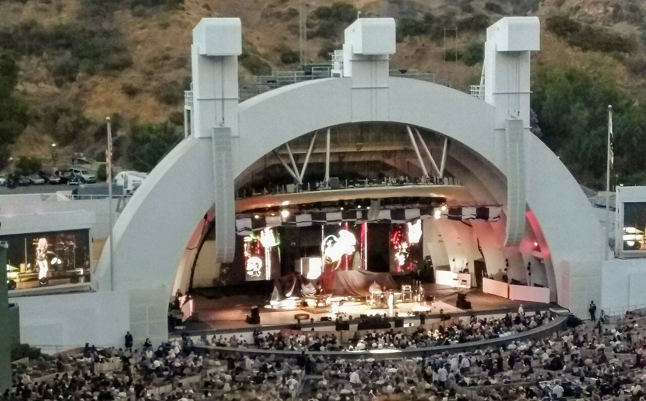 The white arched shell of the Hollywood Bowl lit up for a concert, with a large crowd in the seats in front and green hills behind