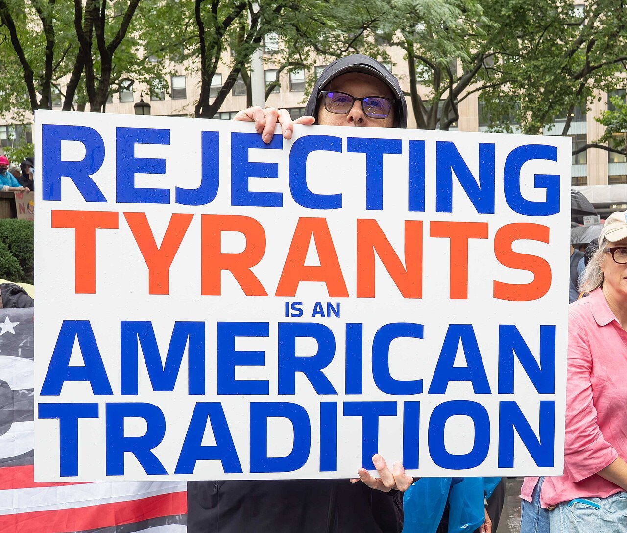 A protester in a cap holds up a large sign reading "Rejecting Tyrants Is an American Tradition" on a city street lined with trees