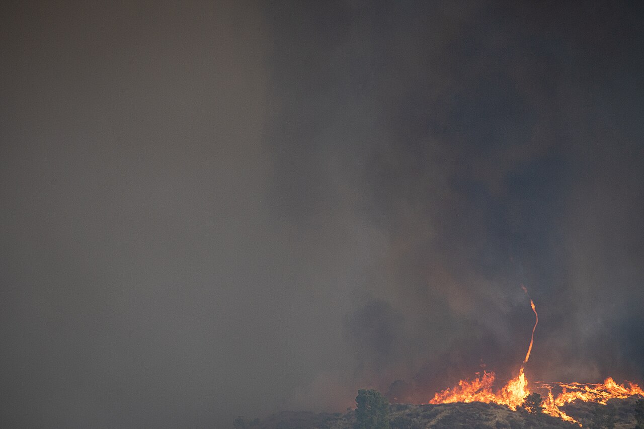 Flames burn along a brush-covered ridge beneath a thick wall of gray smoke, with a thin fire whirl rising from the flames