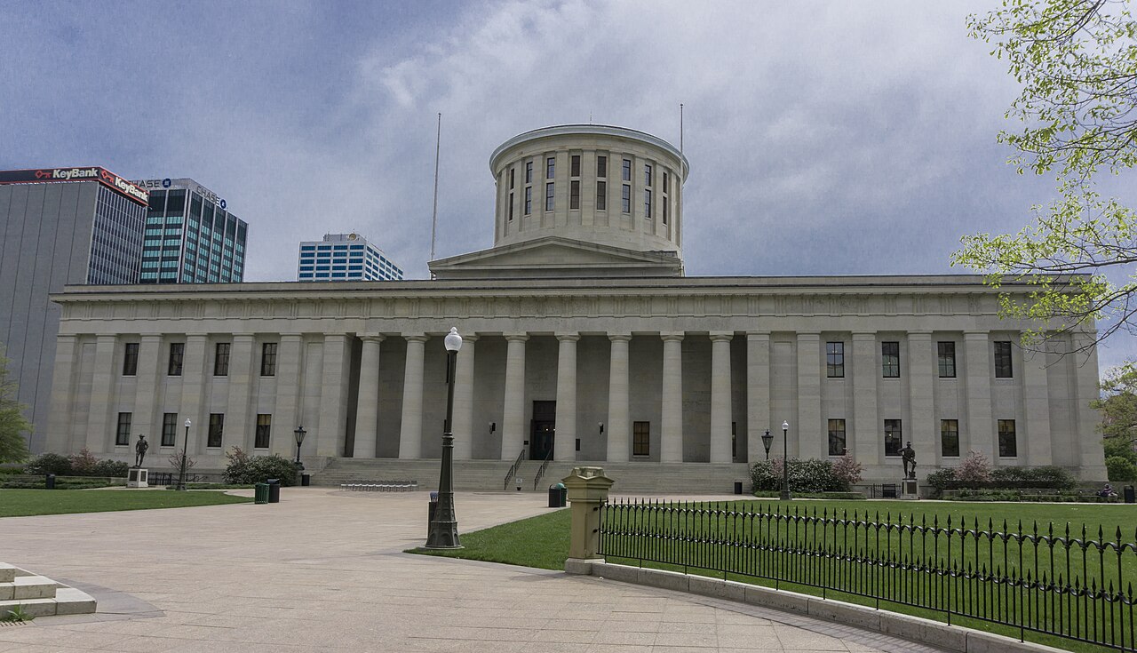 The Ohio Statehouse in Columbus, a pale stone Greek Revival building with a row of columns and a round cupola, seen across a plaza under a cloudy sky