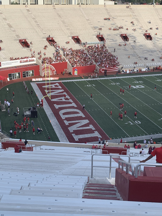 Memorial Stadium in Bloomington, Indiana, seen from the upper stands before a game, with INDIANA painted in the red end zone and players warming up