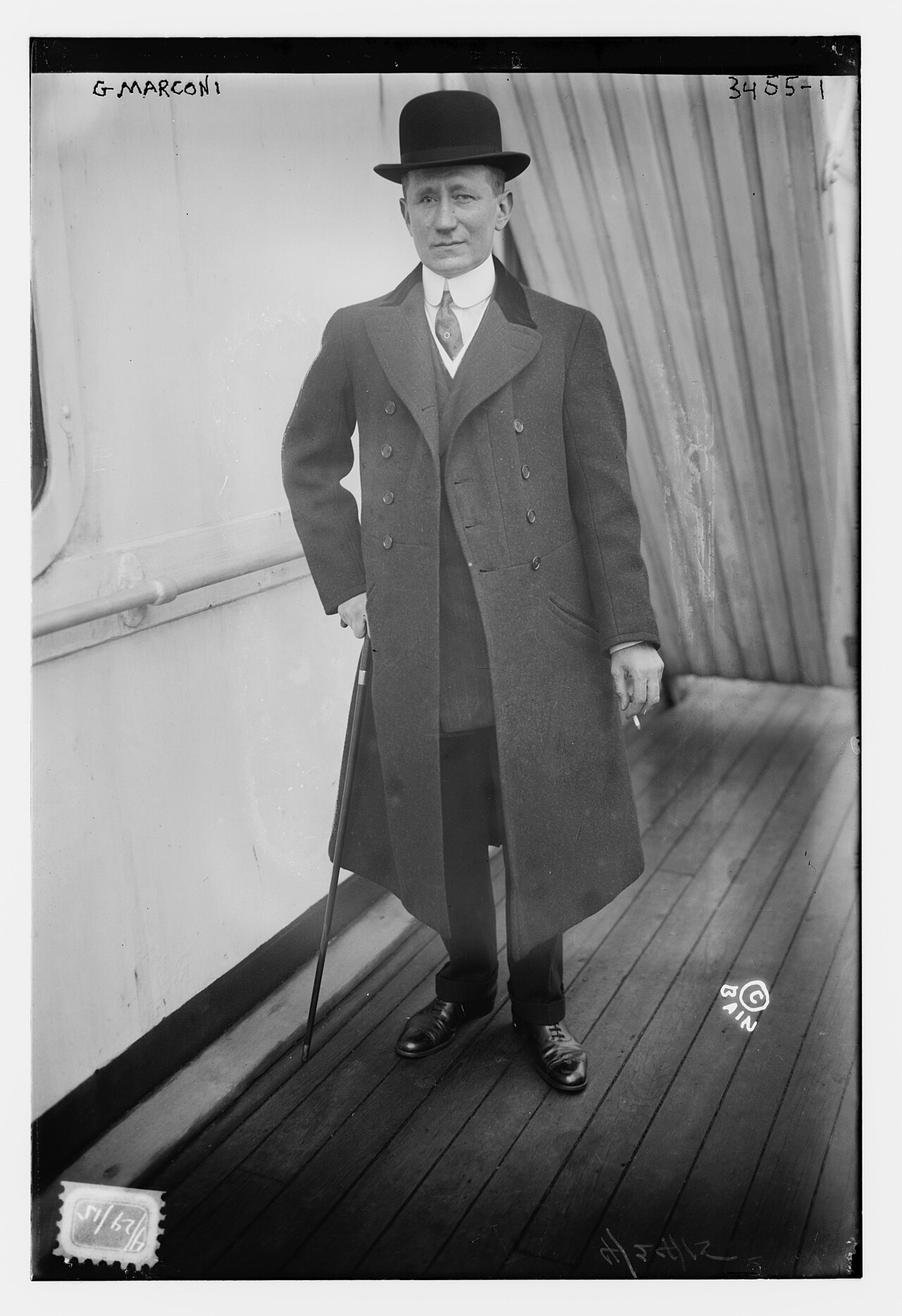 Guglielmo Marconi in a long dark overcoat and bowler hat, holding a cane, standing on a wooden deck
