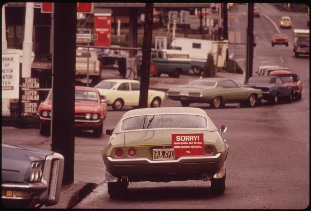 Cars line up along a street at a Shell gas station in 1973, one with a red sign on its back reading 'Sorry! Temporarily out of gas'