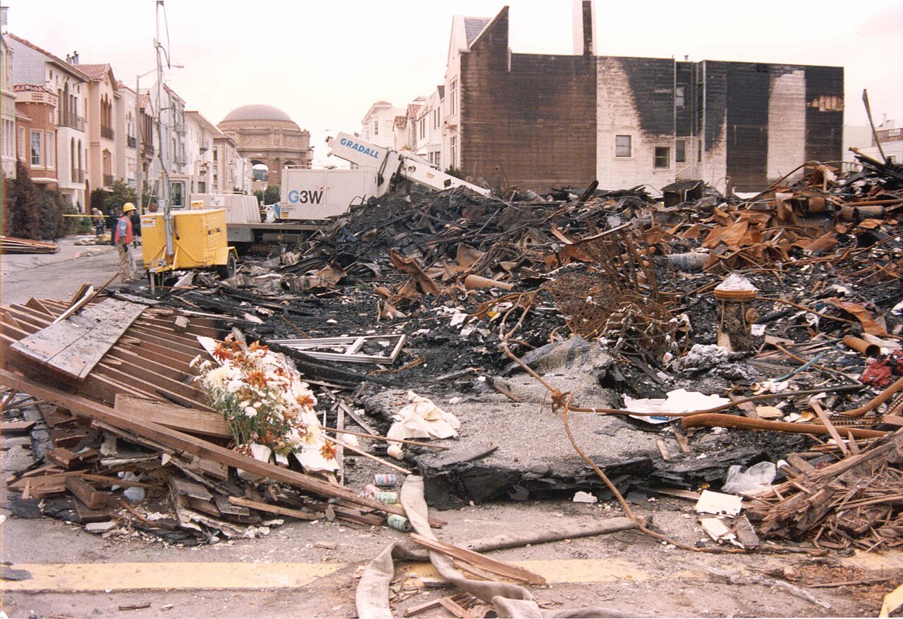 Piles of charred wood and debris on a street lined with damaged buildings, with a yellow construction vehicle and a worker in the background