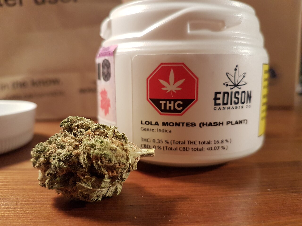 A white plastic jar of legal cannabis labeled Lola Montes by Edison Cannabis Co. next to a dried cannabis bud on a wooden table