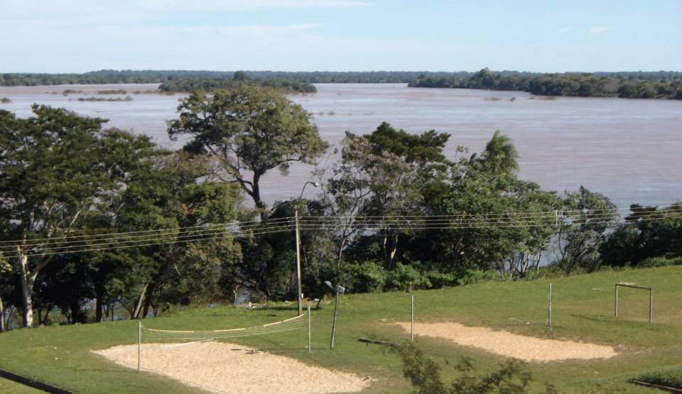 A wide stretch of the Paraná River seen from a grassy riverbank in Ayolas, Paraguay, with trees in the foreground and the far shore on the horizon