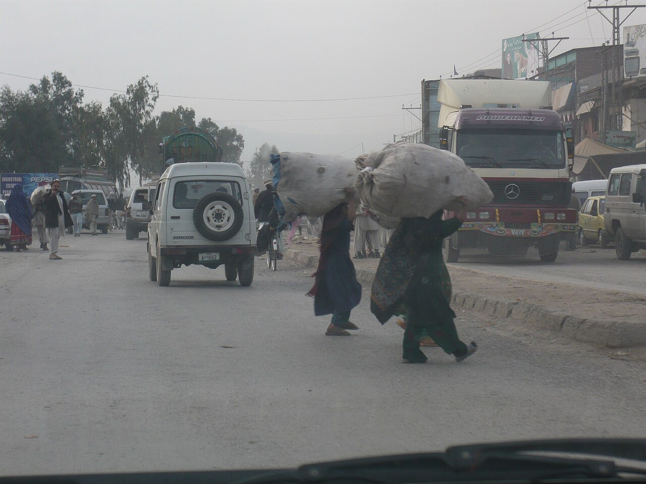 A busy road in Peshawar with a white SUV, a truck and two people carrying large sacks, under a hazy sky