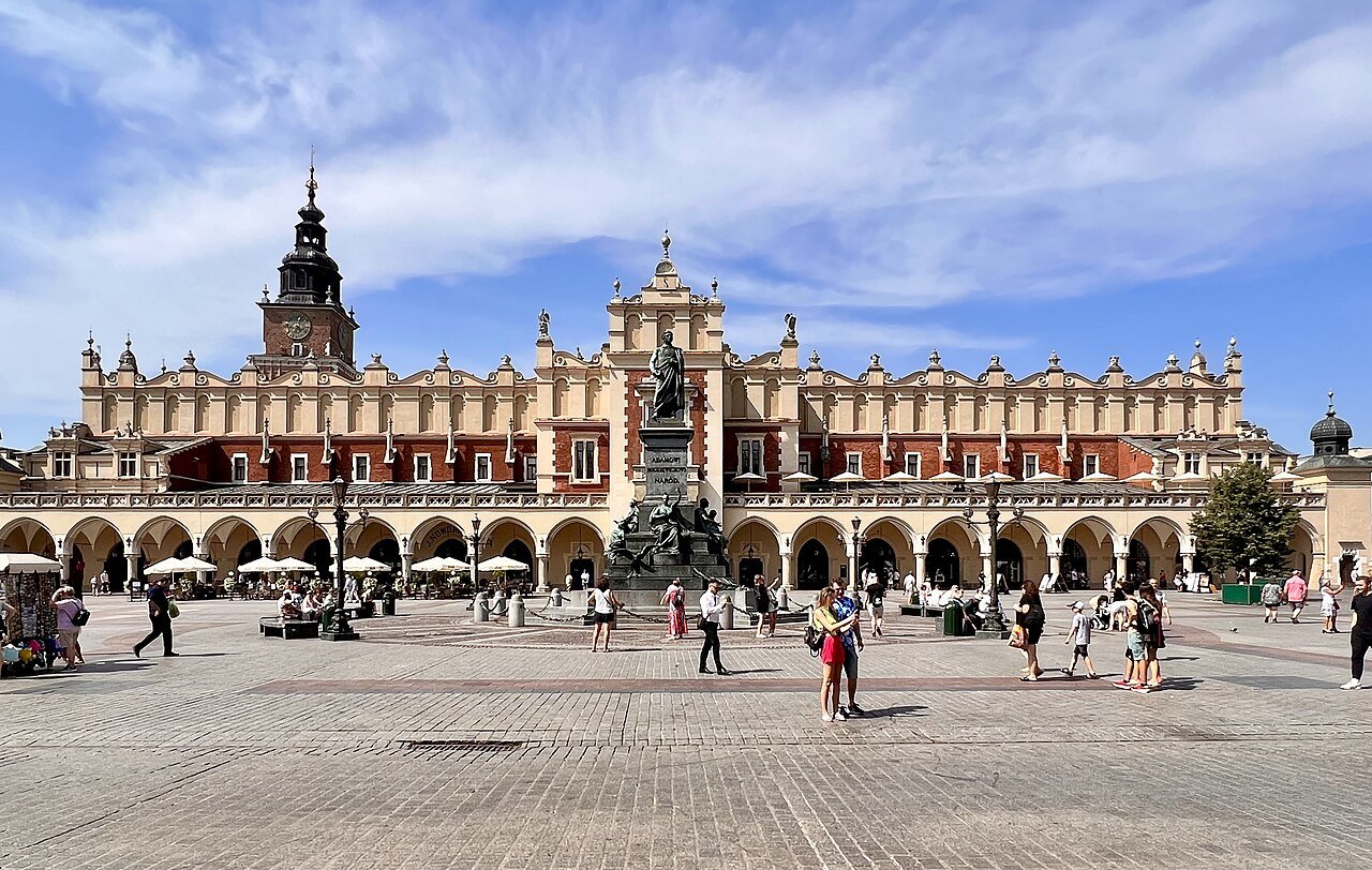 The long Renaissance facade of the Cloth Hall in Kraków's Main Market Square under a blue sky, with people walking across the paved square