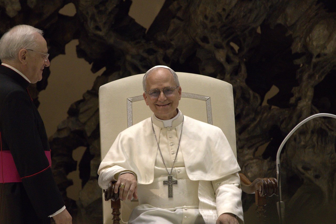 Pope Leo XIV, in white papal robes and a pectoral cross, sits in a white chair during an audience, with a man in black clerical dress partly visible at left