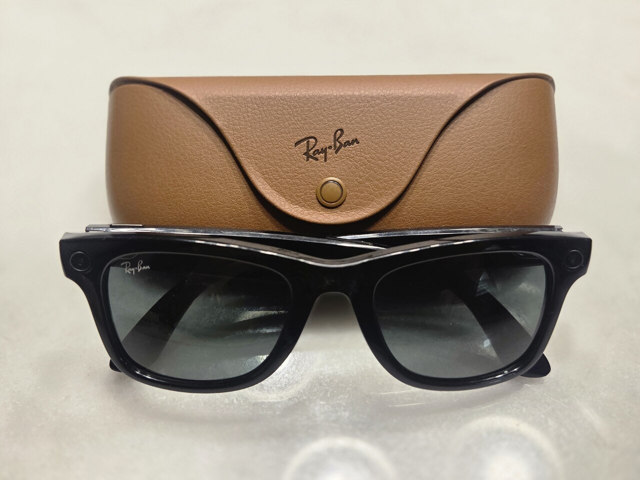 A pair of black Ray-Ban Meta smart glasses with dark lenses resting in front of a tan leather Ray-Ban charging case