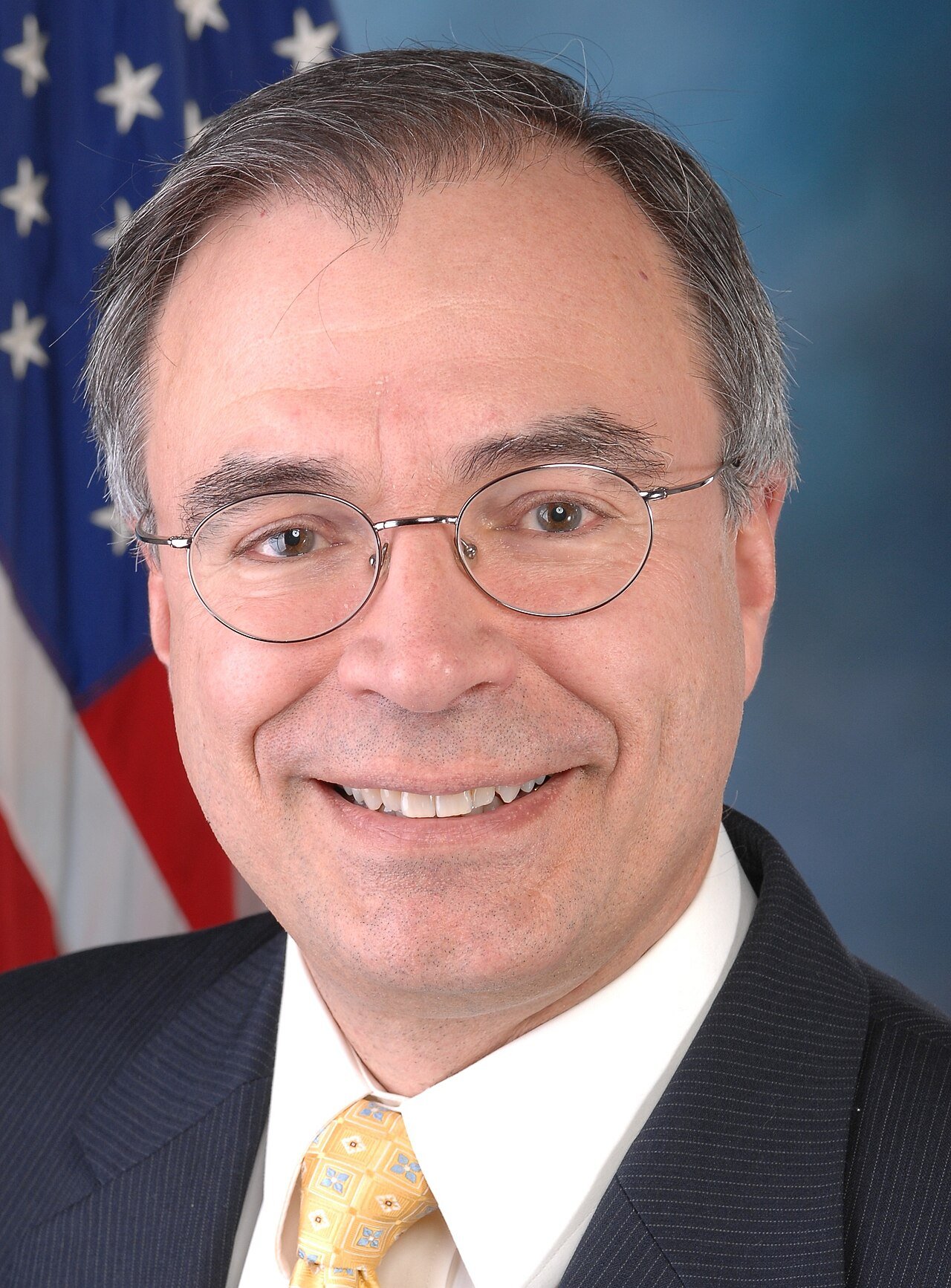 Official portrait of Rep. Andy Harris in a dark suit, white shirt and gold tie, smiling in front of a U.S. flag