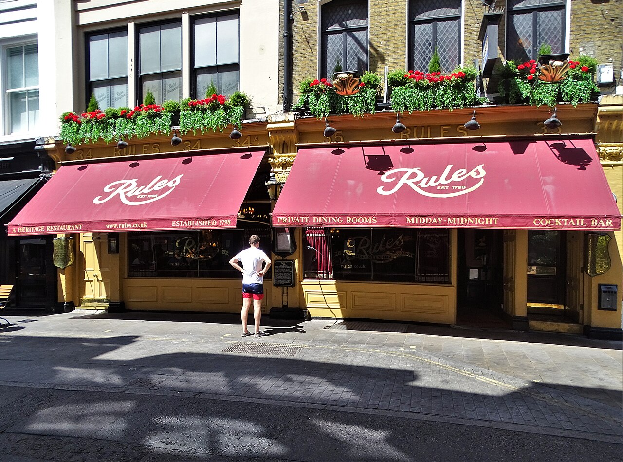 The front of Rules restaurant on a London street, with red awnings, window boxes of red flowers and a man standing on the pavement outside