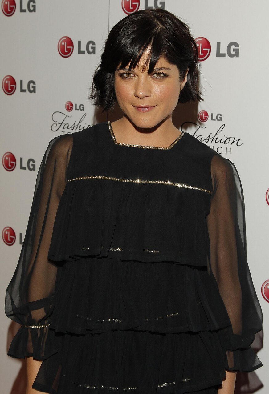 Selma Blair, with a dark bob haircut and a sheer black top, smiles in front of a white backdrop printed with LG logos