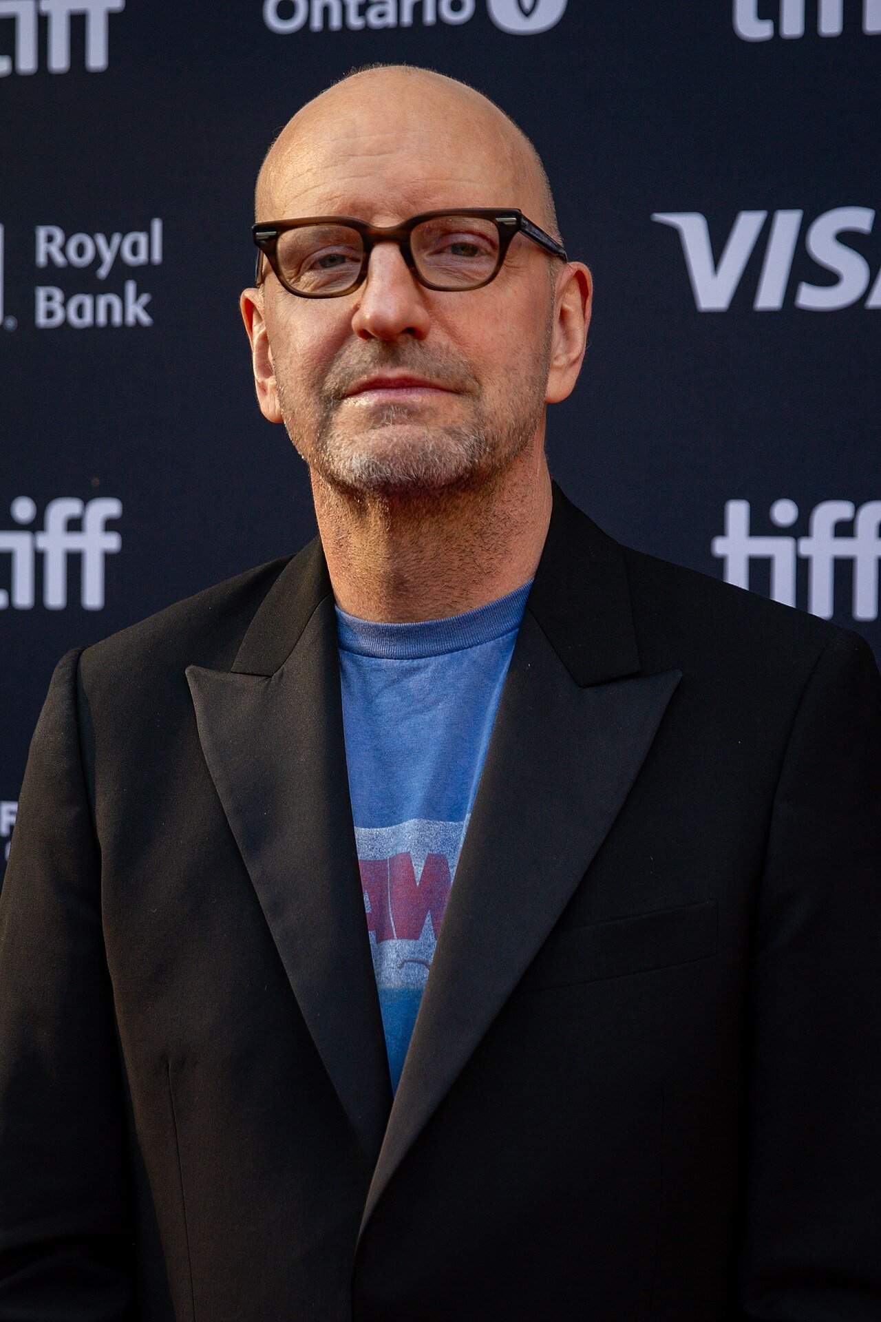 Steven Soderbergh, bald and wearing dark-rimmed glasses, a black blazer and a blue T-shirt, stands in front of a Toronto International Film Festival backdrop
