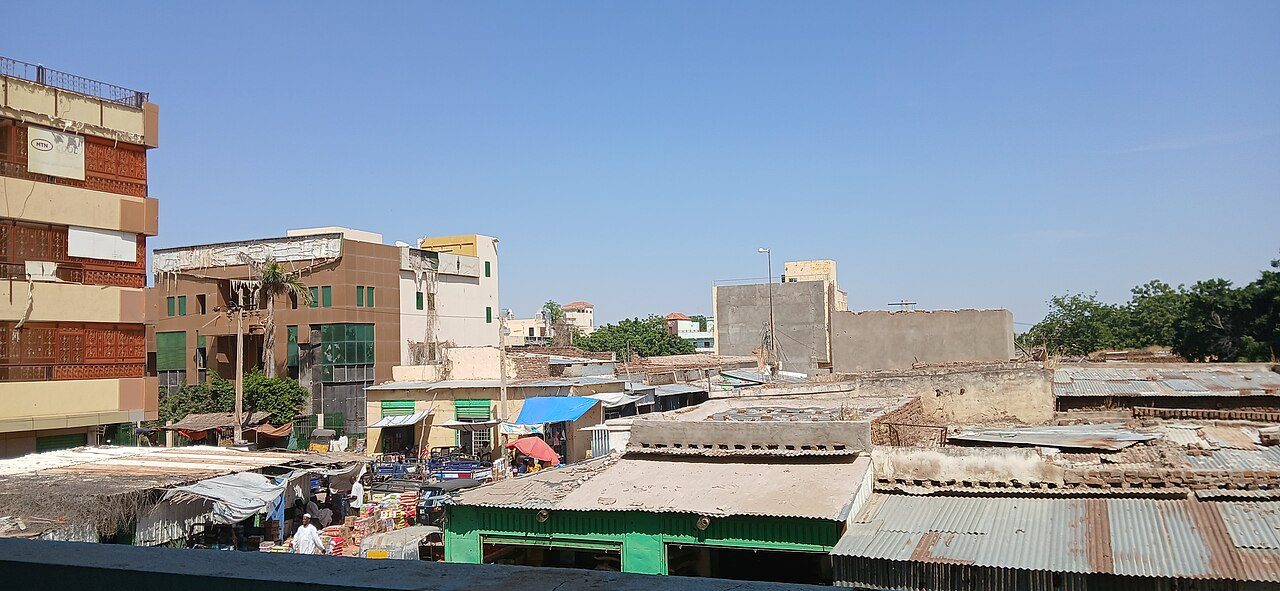 Rooftop view over Nyala's main market in 2025, with rusty corrugated metal roofs, small shops with colored awnings and multistory buildings under a clear blue sky