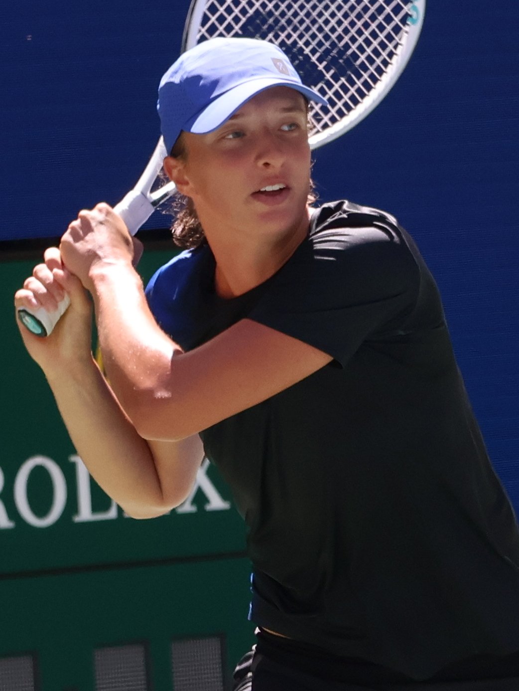 Iga Swiatek in a blue cap and black shirt swings a tennis racket during a practice session