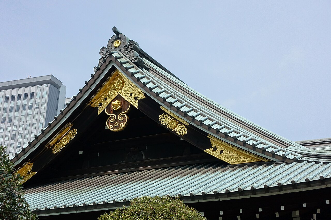 The curved, green-tiled roof of the Yasukuni Shrine main hall in Tokyo, with gold decorations, against a blue sky and a tall building