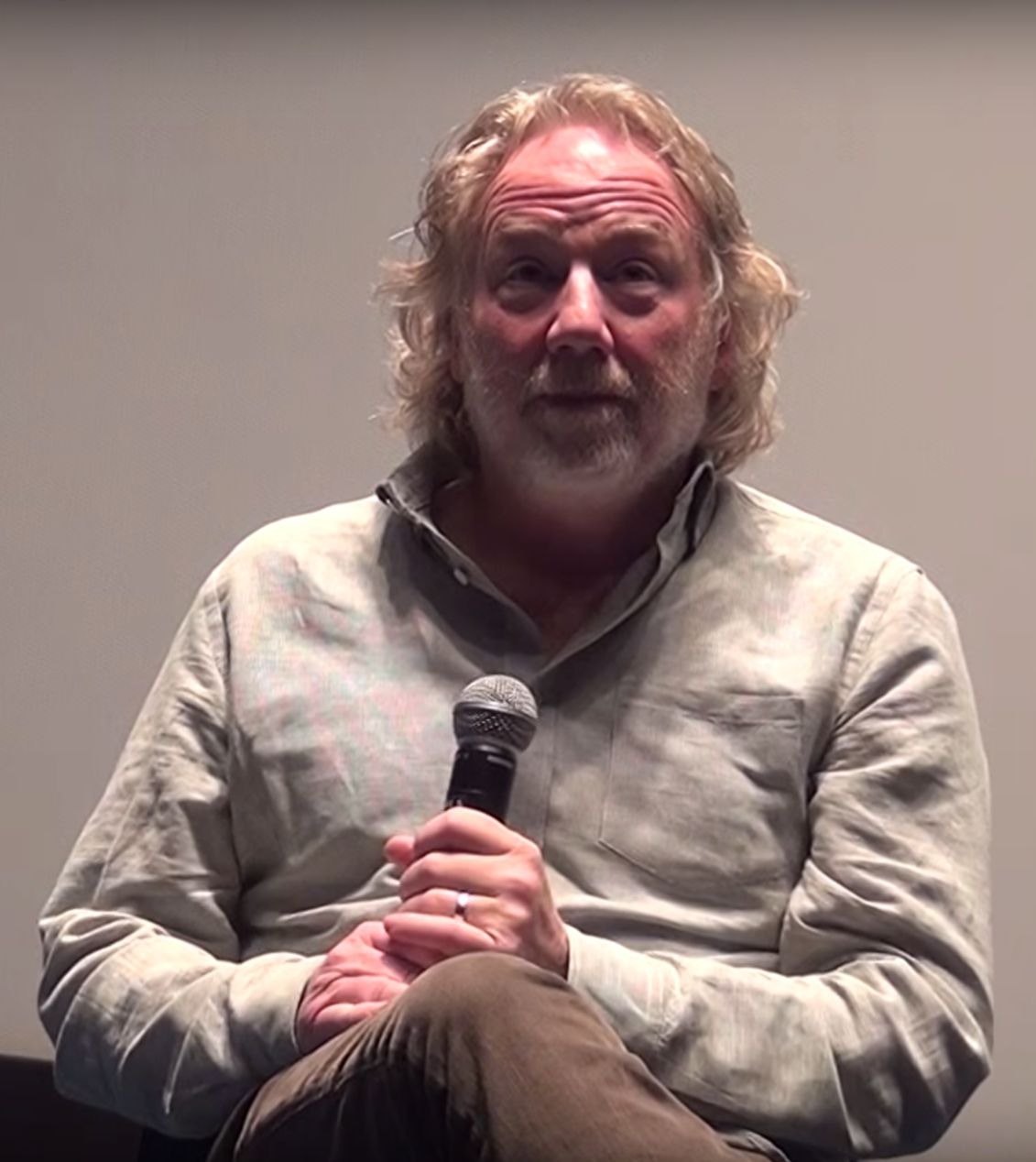 Timothy Busfield, with gray hair and a beard, seated and holding a microphone in front of a pale gray wall