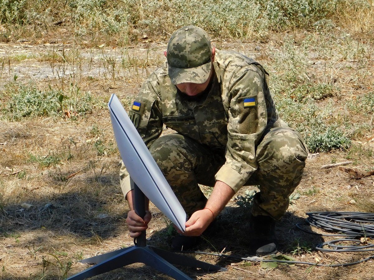 A Ukrainian soldier in camouflage kneels on dry grass while setting up a white Starlink satellite dish on a stand