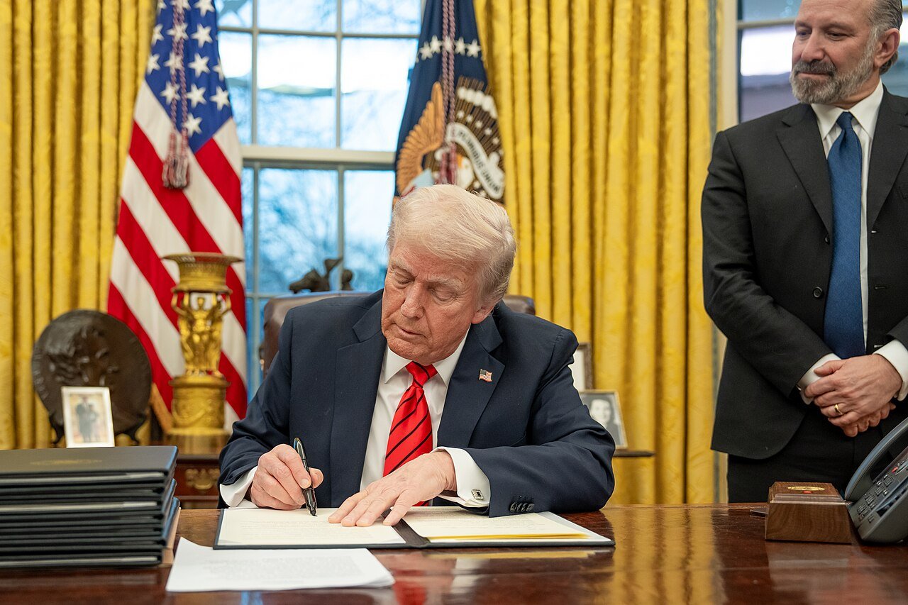 President Donald Trump, seated at the Resolute Desk in the Oval Office, signs a document as a man in a dark suit stands beside him