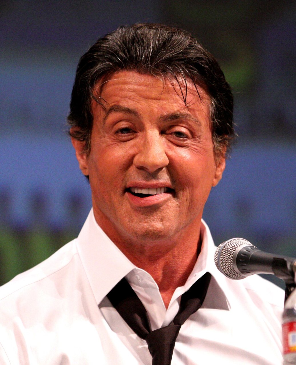 Sylvester Stallone smiling and speaking into a microphone, wearing a white shirt and dark tie, in front of a dark backdrop