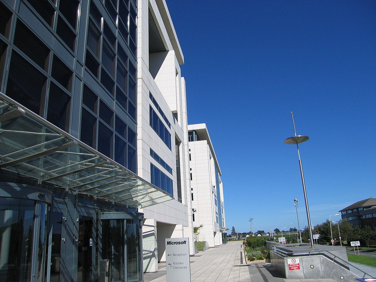 A tall white and glass office building with a Microsoft sign by the entrance pointing to reception and kitchen deliveries, beside a paved walkway and a tall lamp post under a clear blue sky