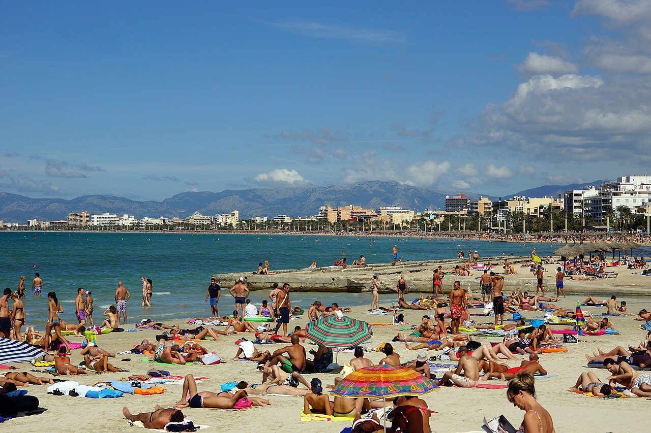 Dozens of sunbathers lie on towels and sit under striped umbrellas on a crowded sandy beach beside turquoise water, with hotels, palm trees and mountains along the curving shoreline behind them