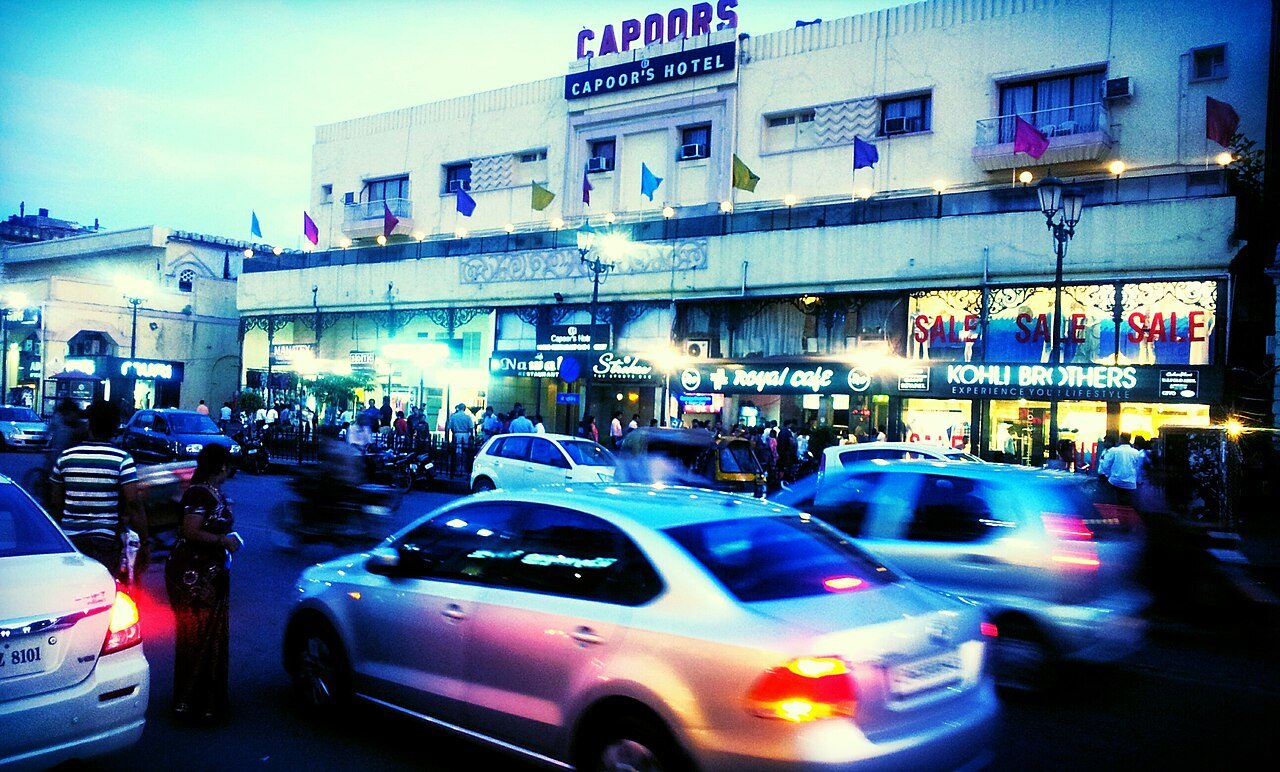 Evening traffic passes lit shops and a hotel on a busy street in Hazratganj, Lucknow