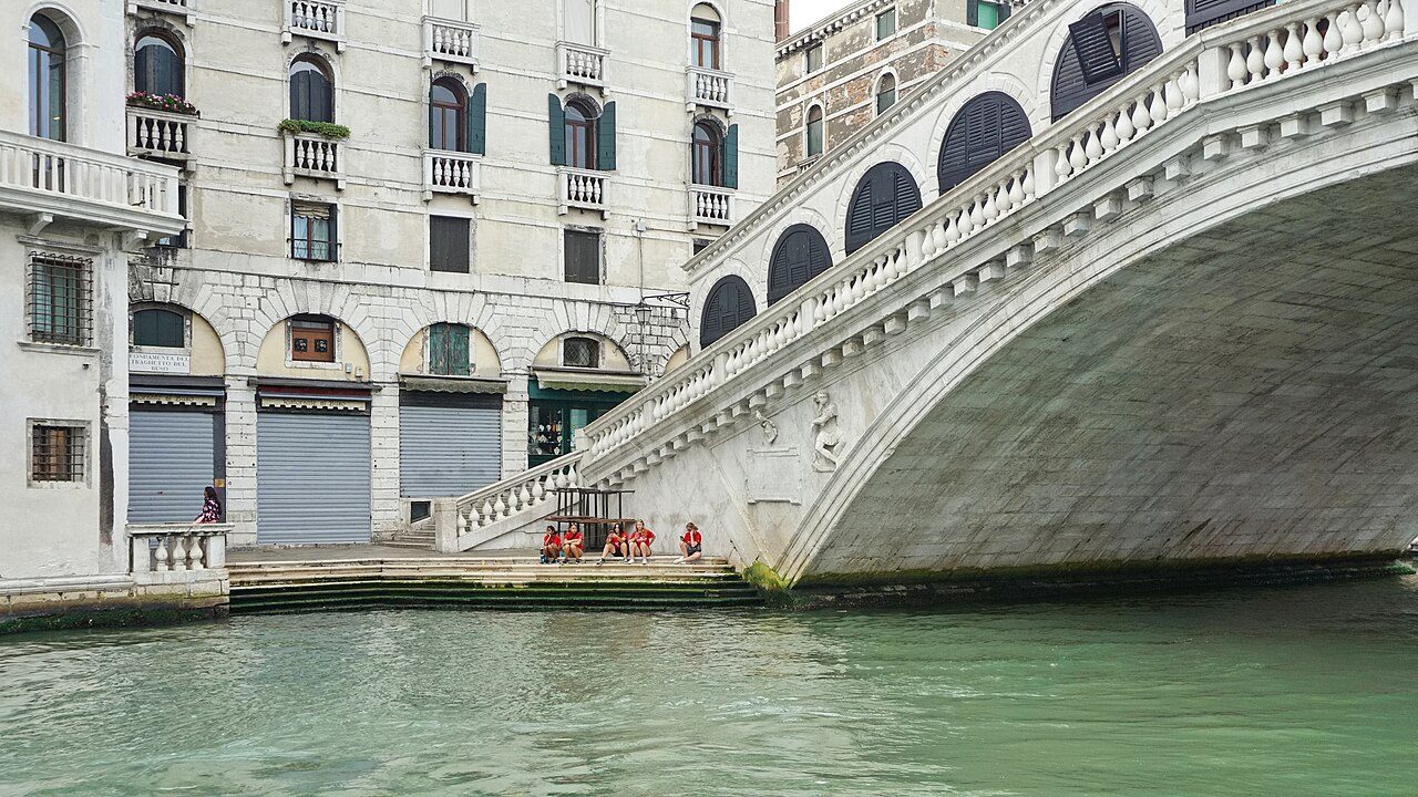 The white stone arch of Venice's Rialto Bridge over the green water of the Grand Canal, next to a pale stone building with arched windows