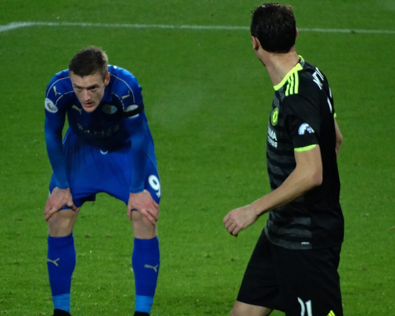 Jamie Vardy in a blue Leicester City kit bends forward with his hands on his knees during a match, as a player in black walks past