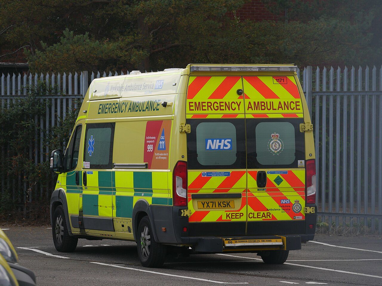 A yellow and green Yorkshire Ambulance Service emergency ambulance, with NHS and Keep Clear markings on its back doors, parked beside a tall metal fence