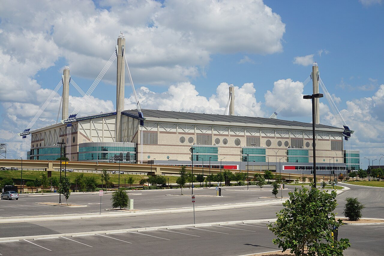 A large white domed stadium with four tall corner towers stands beyond an empty parking lot under a cloudy blue sky