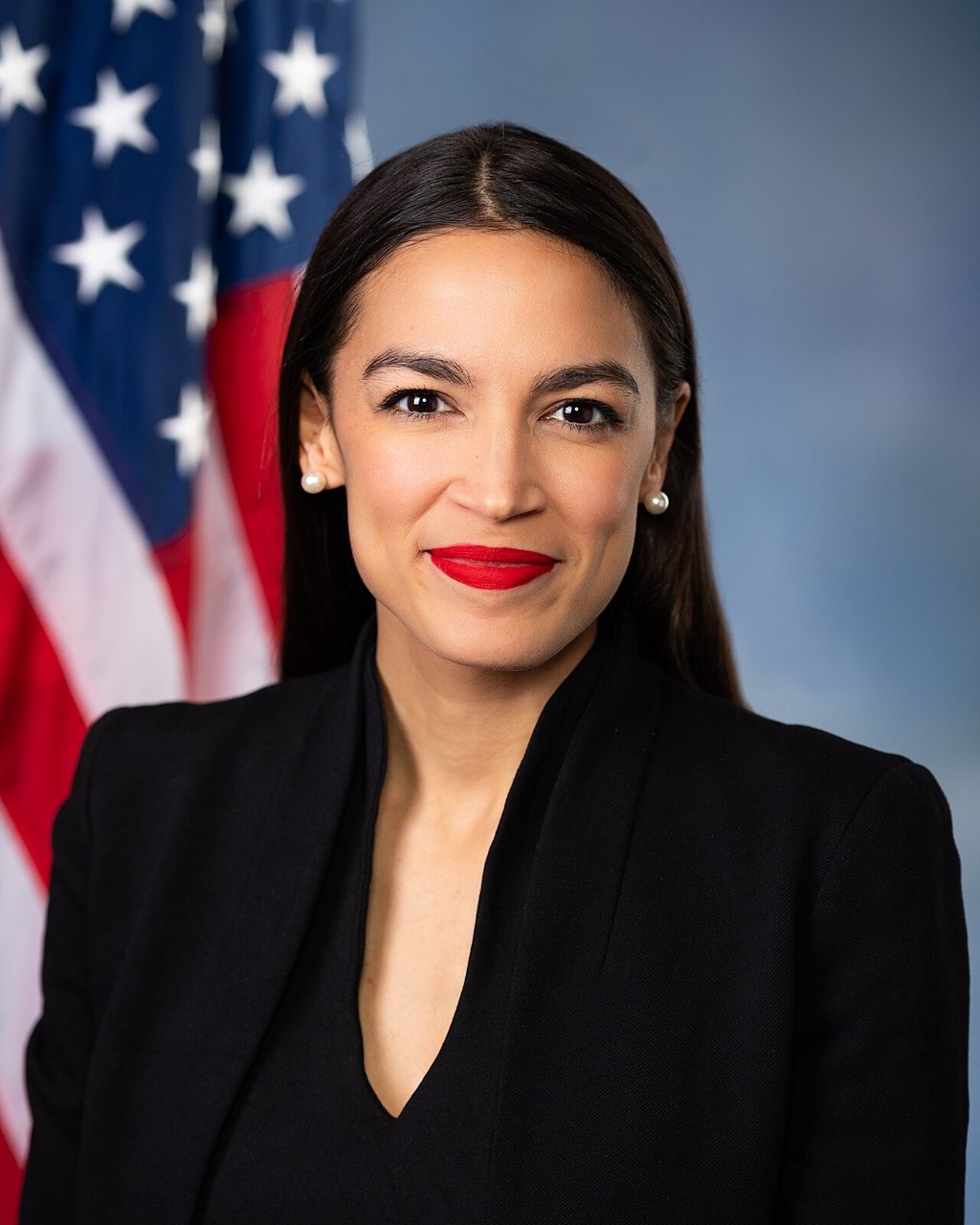 Alexandria Ocasio-Cortez in a black jacket, smiling, in front of a US flag