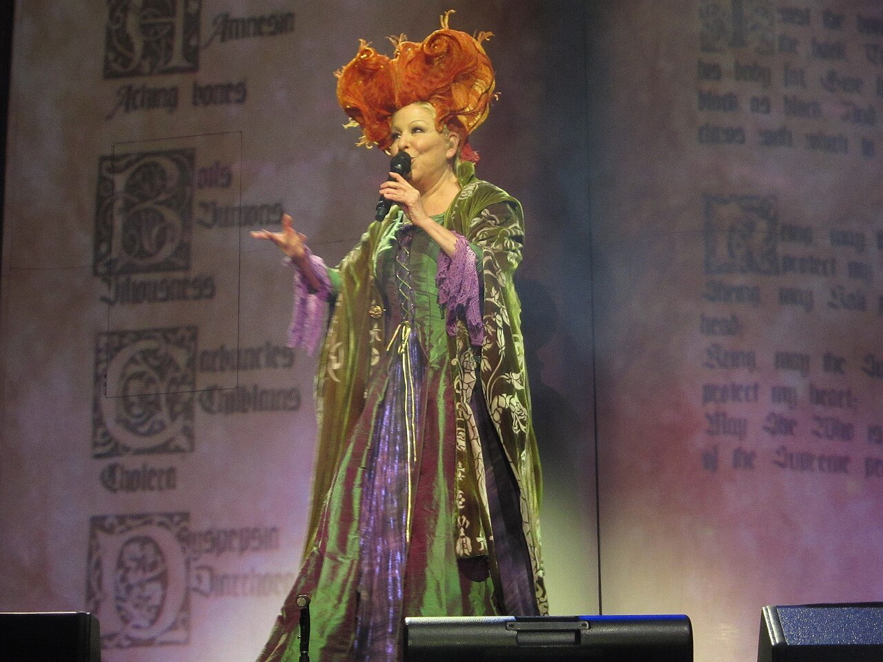 Bette Midler in a flowing green and purple costume and red wig, singing on stage
