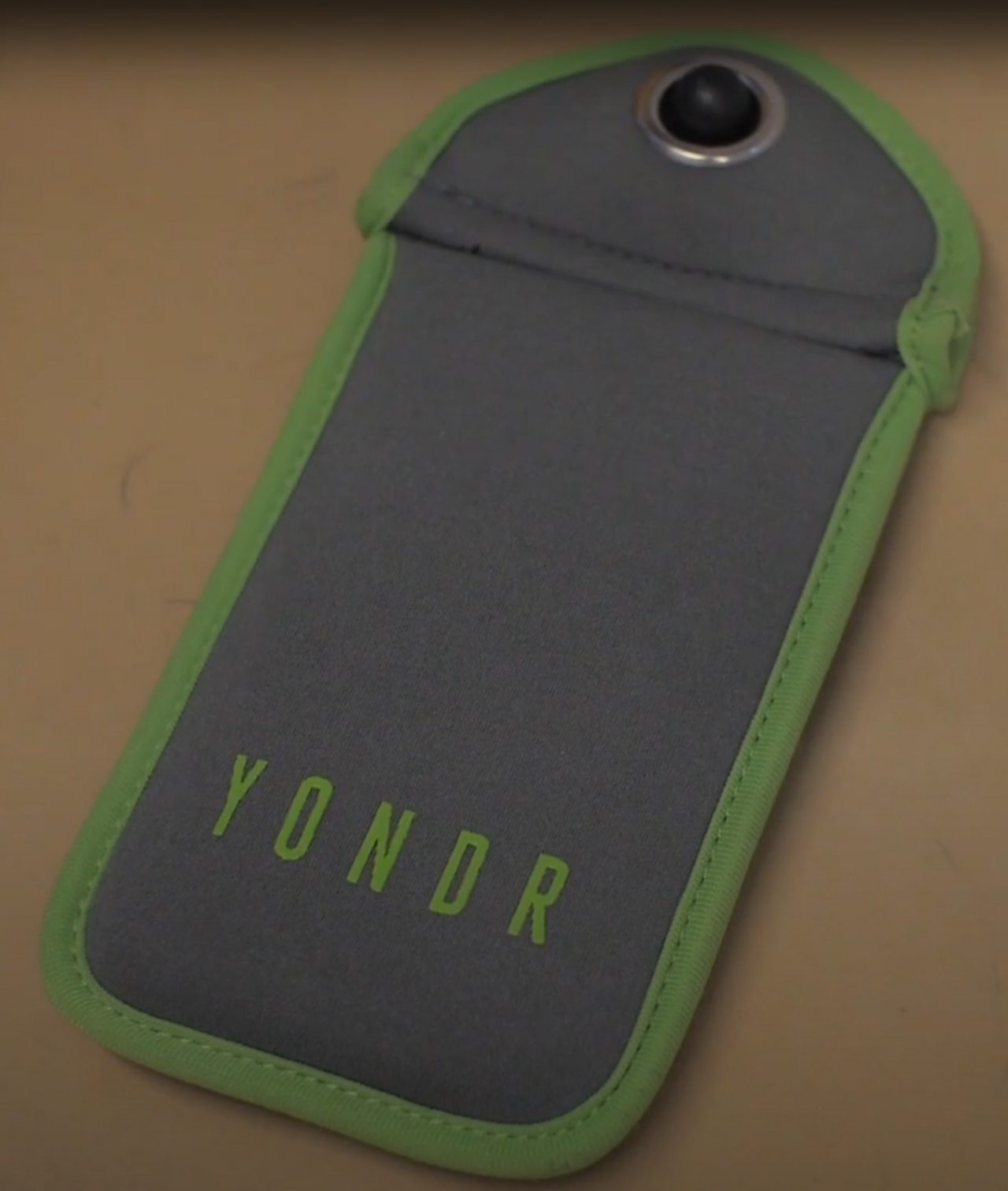 A gray fabric phone pouch with a green rim and a round metal lock button at the top lies on a wooden surface, with the word YONDR printed on it
