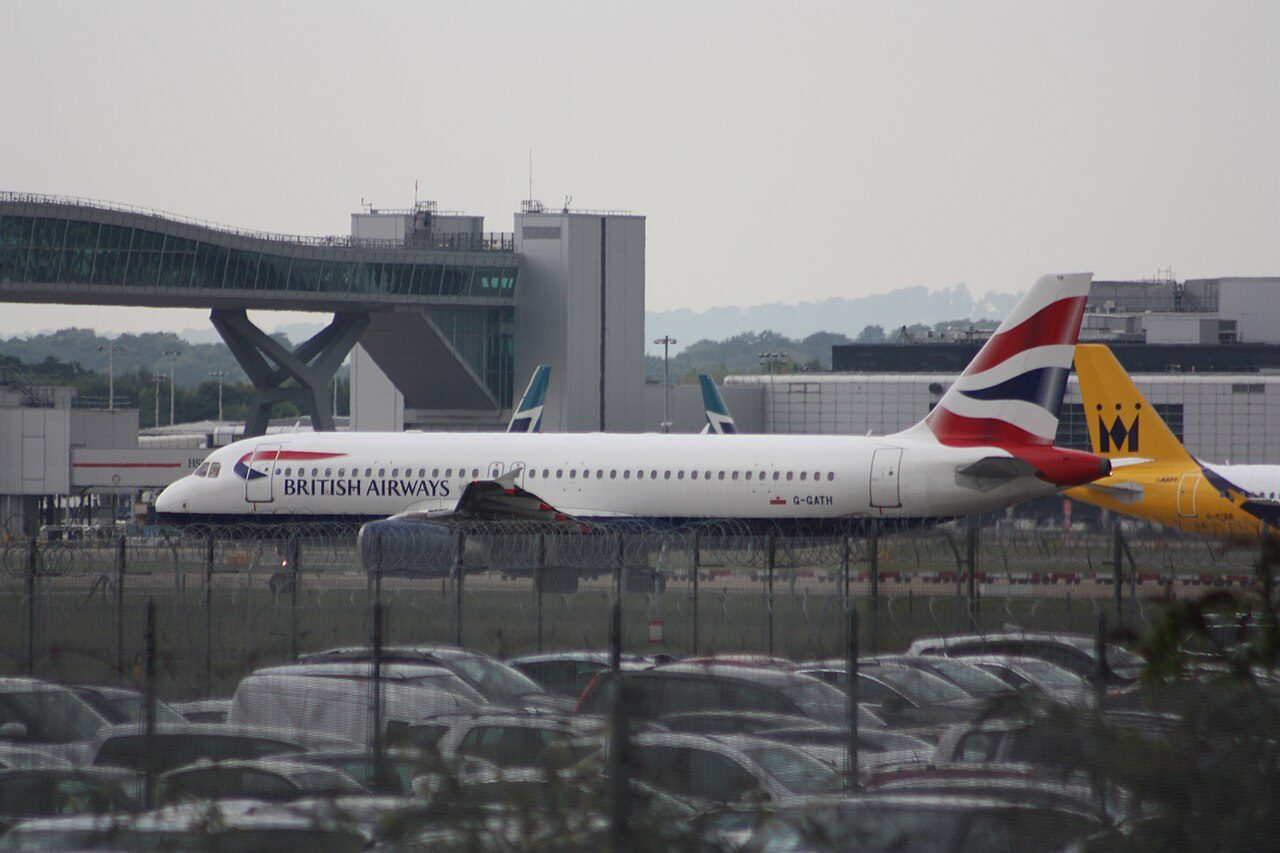 A white British Airways jet with a red, white and blue tail sits on the tarmac at London Gatwick Airport, with a terminal building and parked cars in front