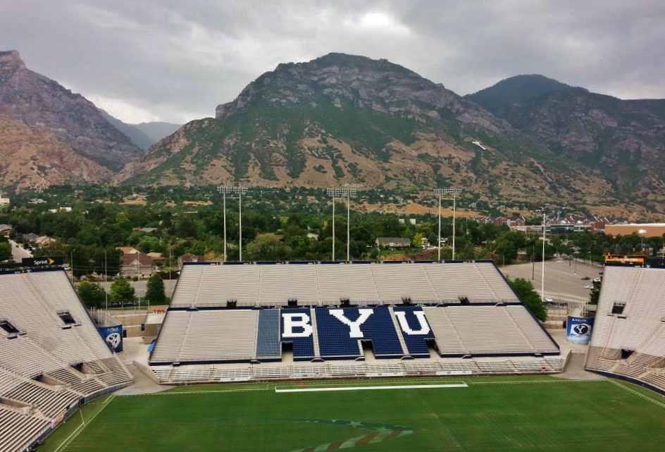 An empty football stadium with blue BYU lettering in the stands and green field, with tall mountains rising behind it under a cloudy sky