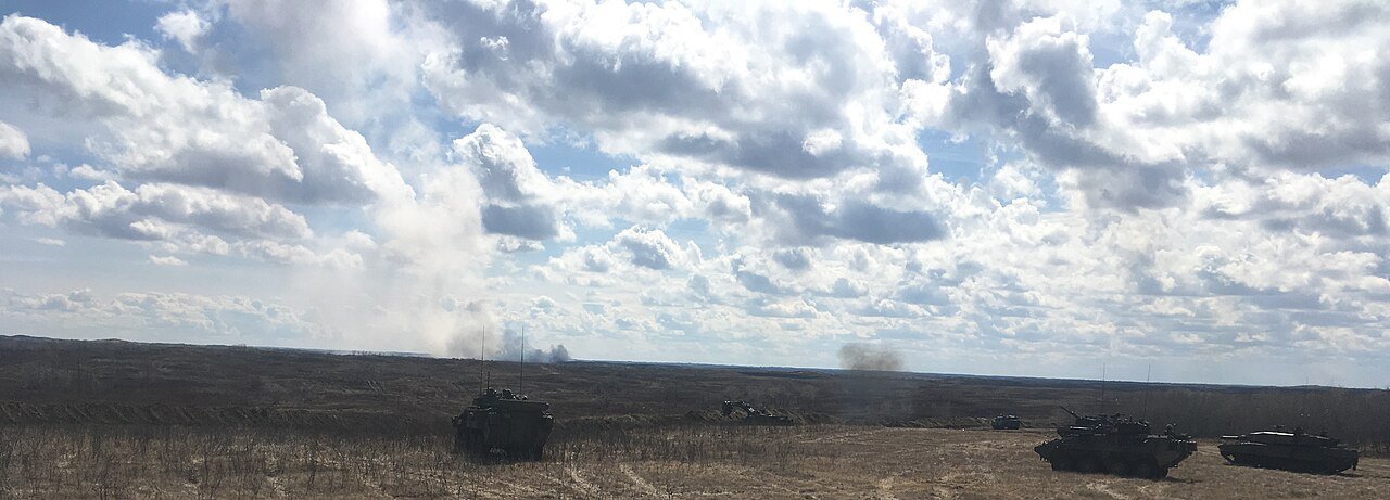 Armoured vehicles spread across a flat, dry training field under a cloudy sky as smoke rises in the distance