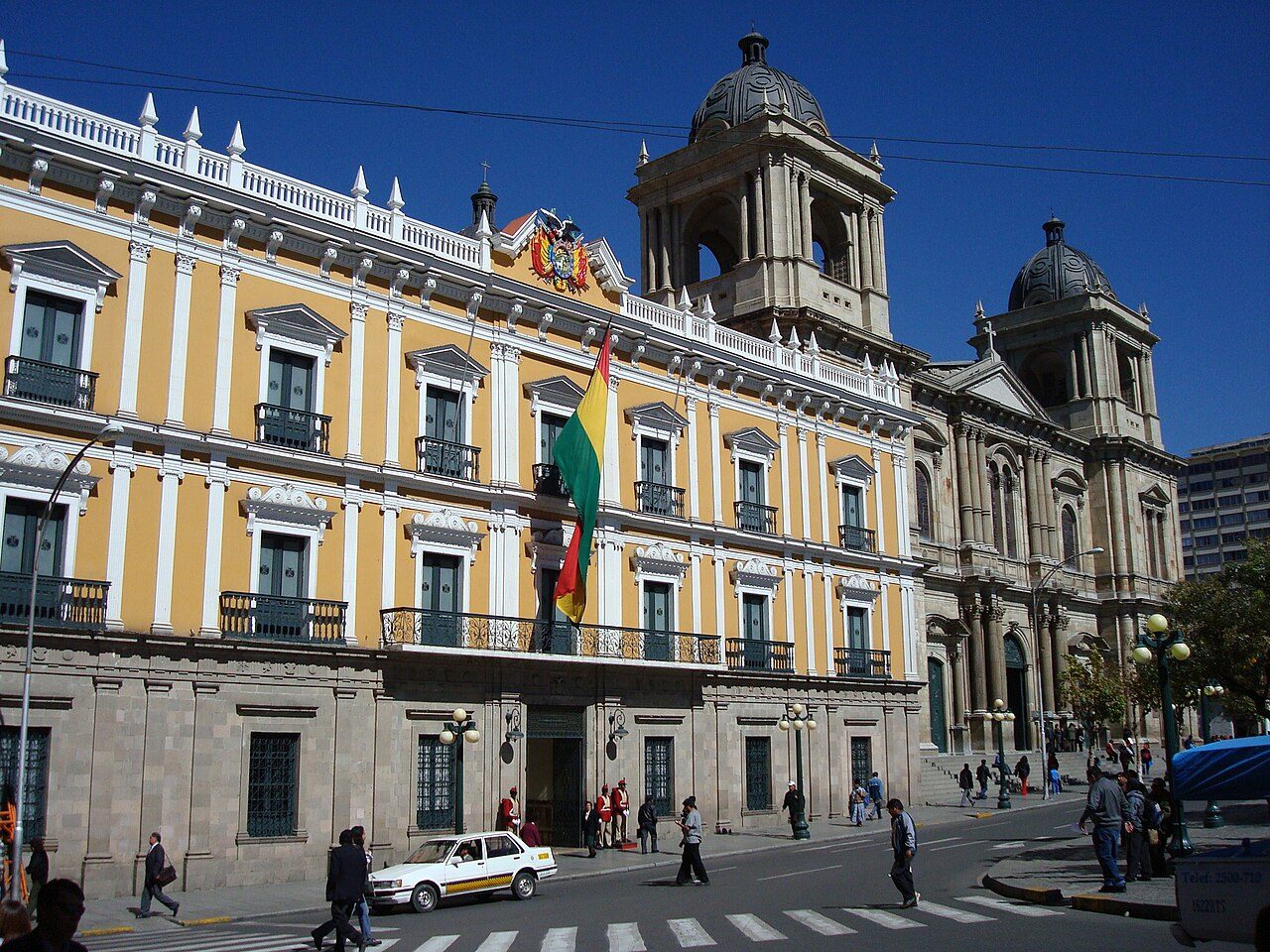 A pale yellow colonial-style government building with white columns and balconies stands beside a domed stone cathedral on a sunny plaza, with a few cars and people on the street in front