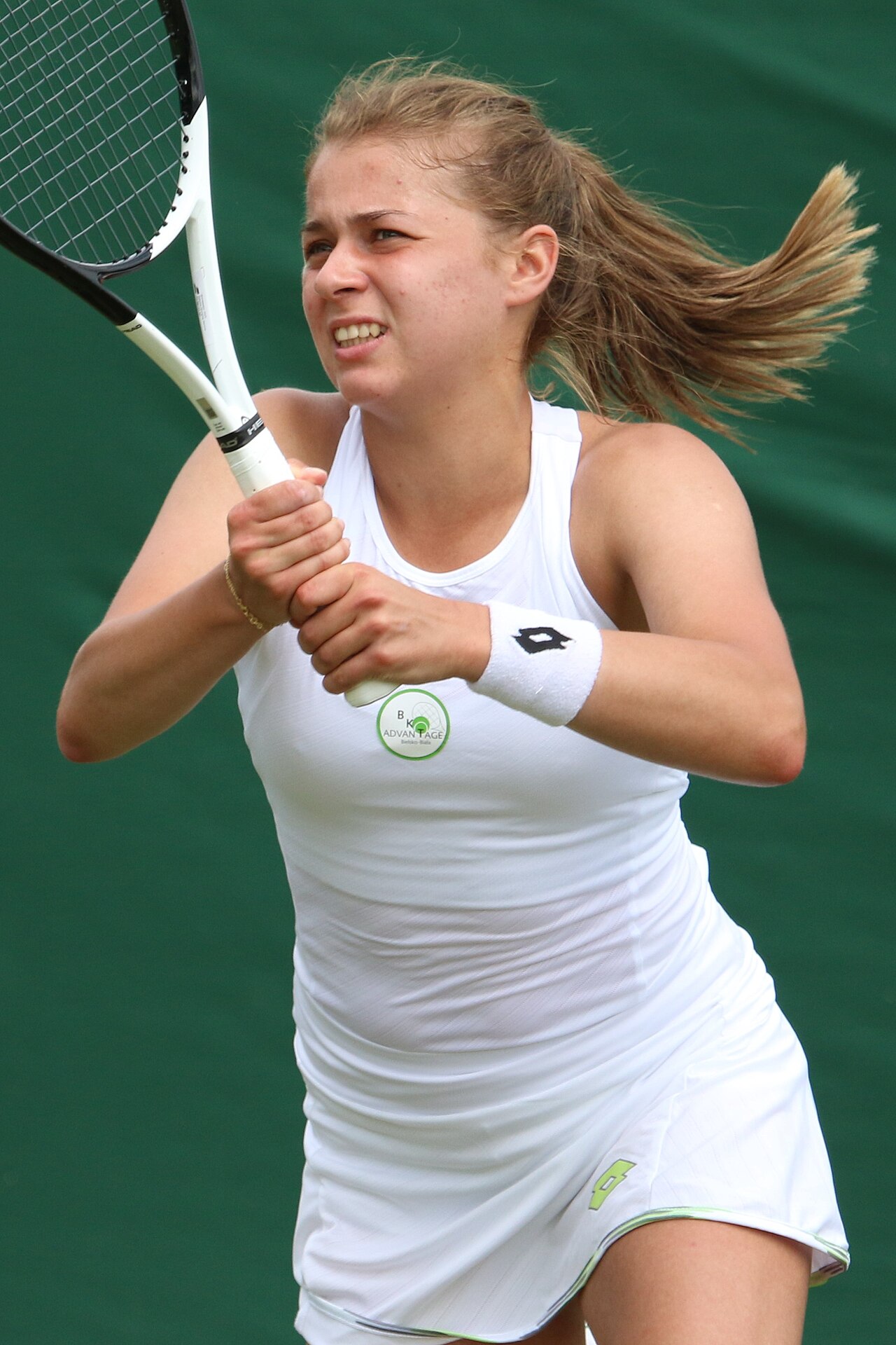 A tennis player in a white dress swings a racket with both hands on a grass court in front of a green backdrop