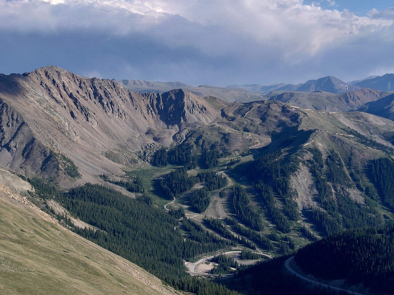 Rocky mountain slopes with green forest and gray rock under a partly cloudy sky, with ski runs cut through the trees on the north side of Arapahoe Basin