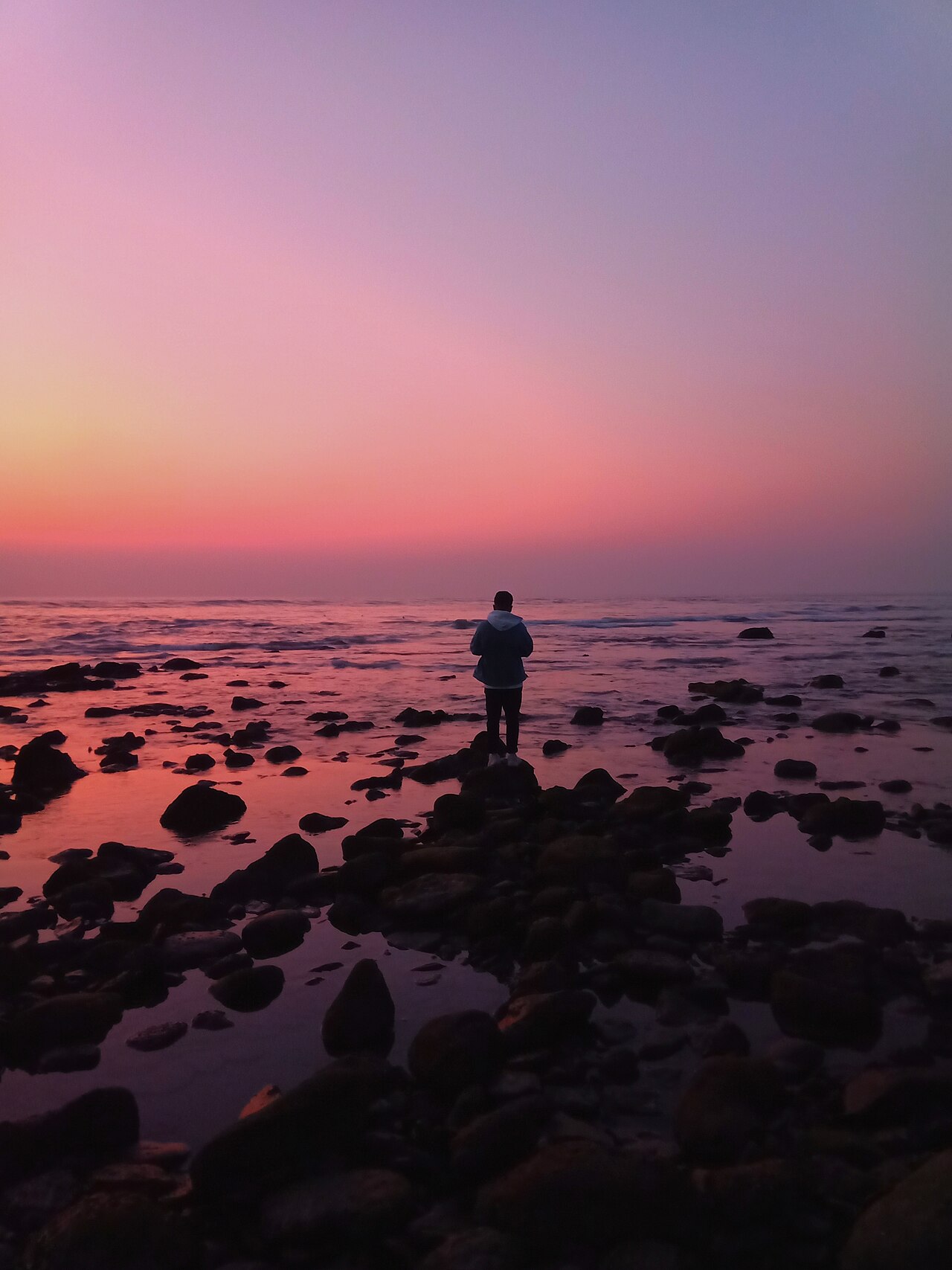 A lone person stands silhouetted on rocks at the edge of the sea under a pink and purple sunset sky