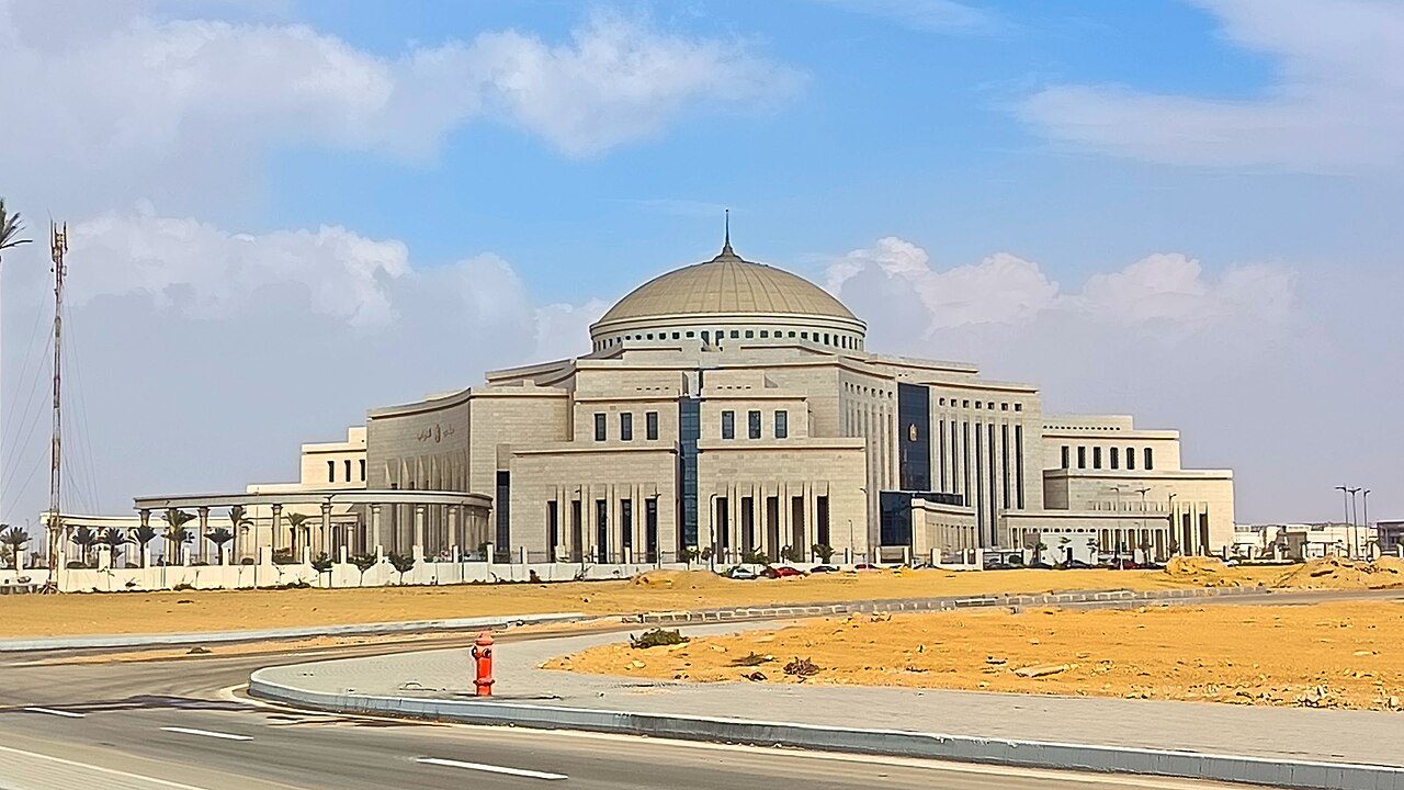 A large pale stone building with a central dome and rows of columns stands behind a curving road and stretches of sand under a blue sky with scattered clouds