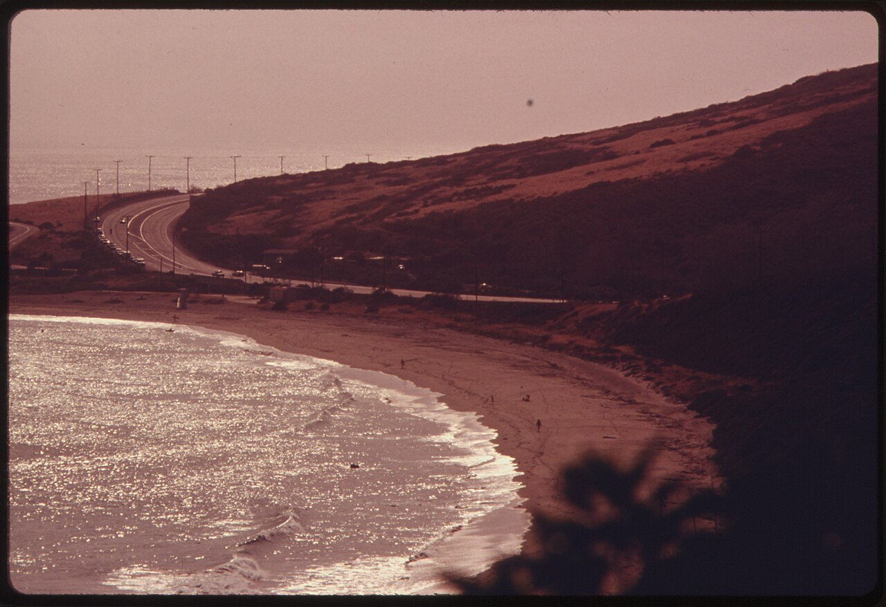 An old reddish-toned photo of a two-lane highway curving along a hillside above a sandy beach and breaking waves
