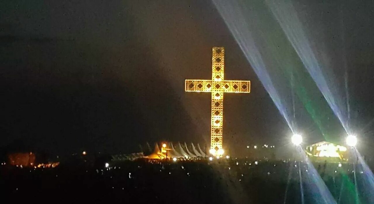A large illuminated cross stands on a dark hillside at night as spotlights sweep the sky