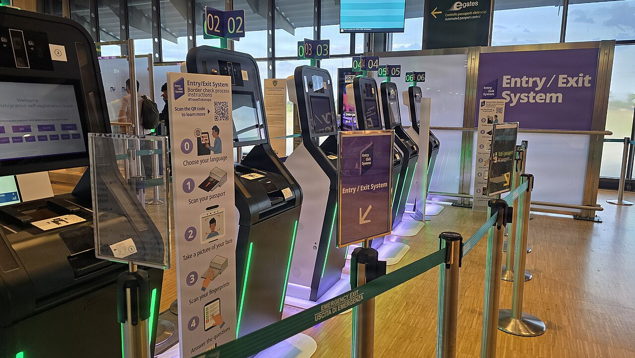 A row of self-service kiosks with screens and a purple Entry/Exit System sign in an airport hall, with queue barriers in front