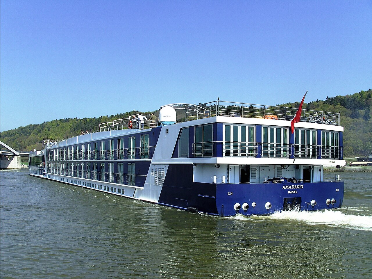 A long blue and white river cruise ship sails on a wide river past green wooded hills