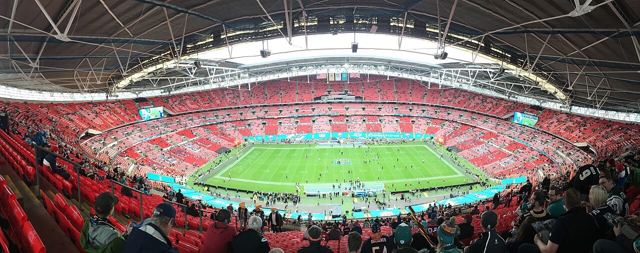 A panoramic view inside a large stadium with red seats and a curved roof, looking down at an American football field before a game, with fans in the foreground